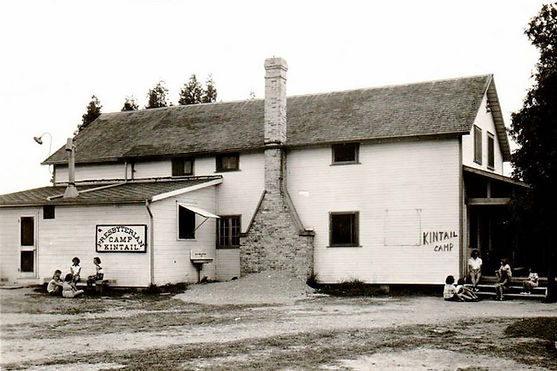 A black and white image of Haromny House. It is a white sided building and a big chimney. There are a few people sitting outside of it.