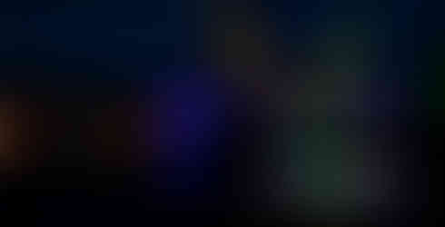 The words DUNHAM MASSEY are lit in multi-colours and are in front of a building lit in blue with yellow lasers in the background. The building is next to water and it is dark.