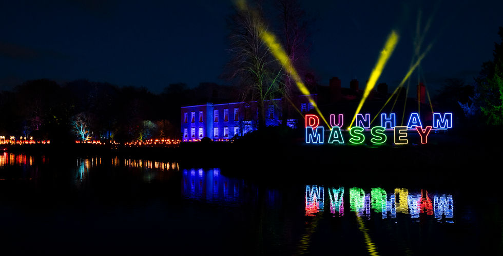 The words DUNHAM MASSEY are lit in multi-colours and are in front of a building lit in blue with yellow lasers in the background. The building is next to water and it is dark.