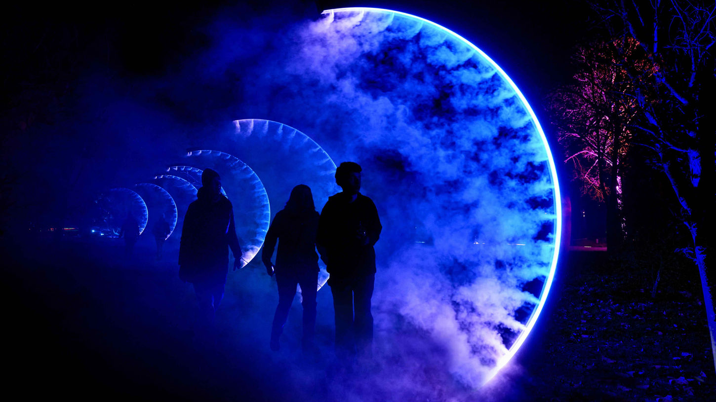A row of curved arches emitting blue smoke. Figures are silhouetted underneath. It is dark with pink and blue lighting.