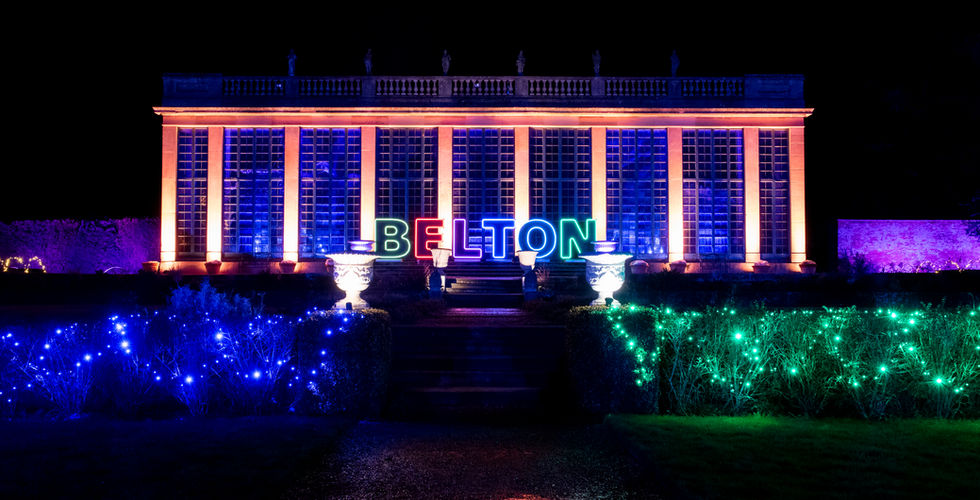 BELTON written in multicoloured lights sits in front of a building made of columns list in orange. There are bushes in the foreground lit in green, blue and red.