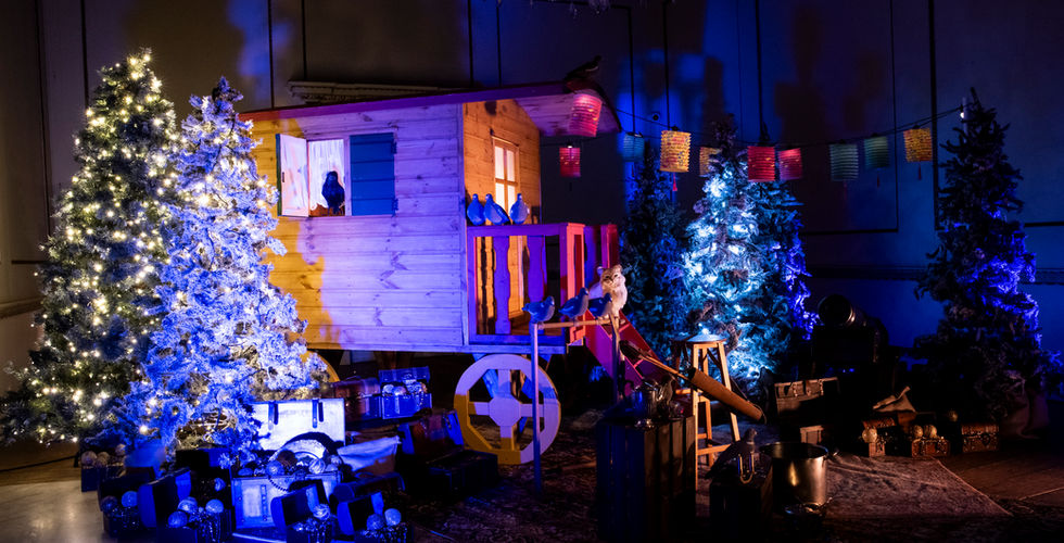 A traditional wooden travelling caravan lit in various colours surrounded by Christmas trees. A cat sits on the top of the steps that lead to the door to the caravan.