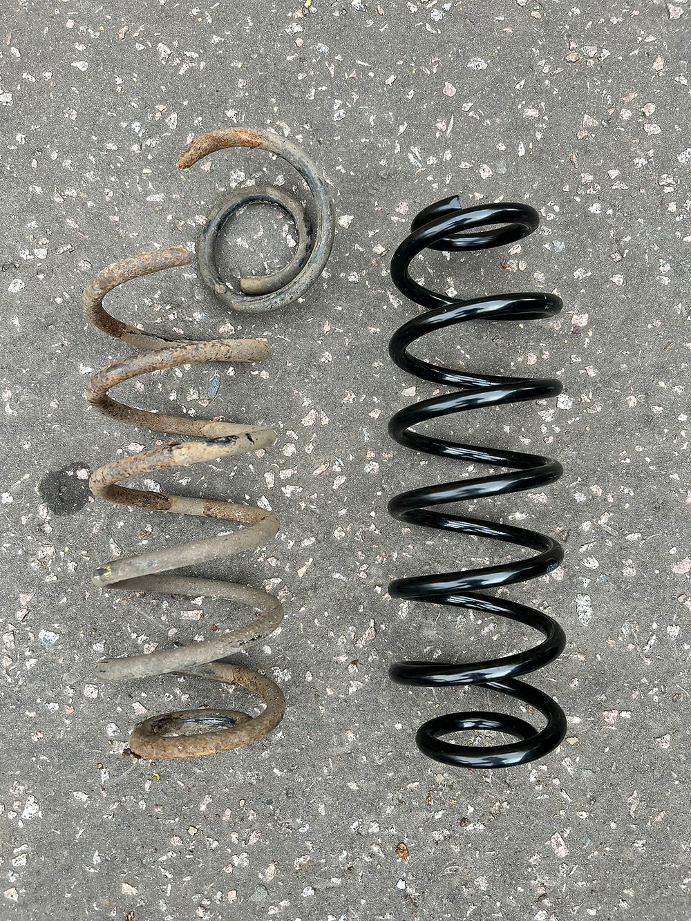 Side-by-side comparison of a snapped, rusty car suspension coil spring next to a brand-new, high-quality replacement spring, professionally fitted during a mobile mechanical repair service.
