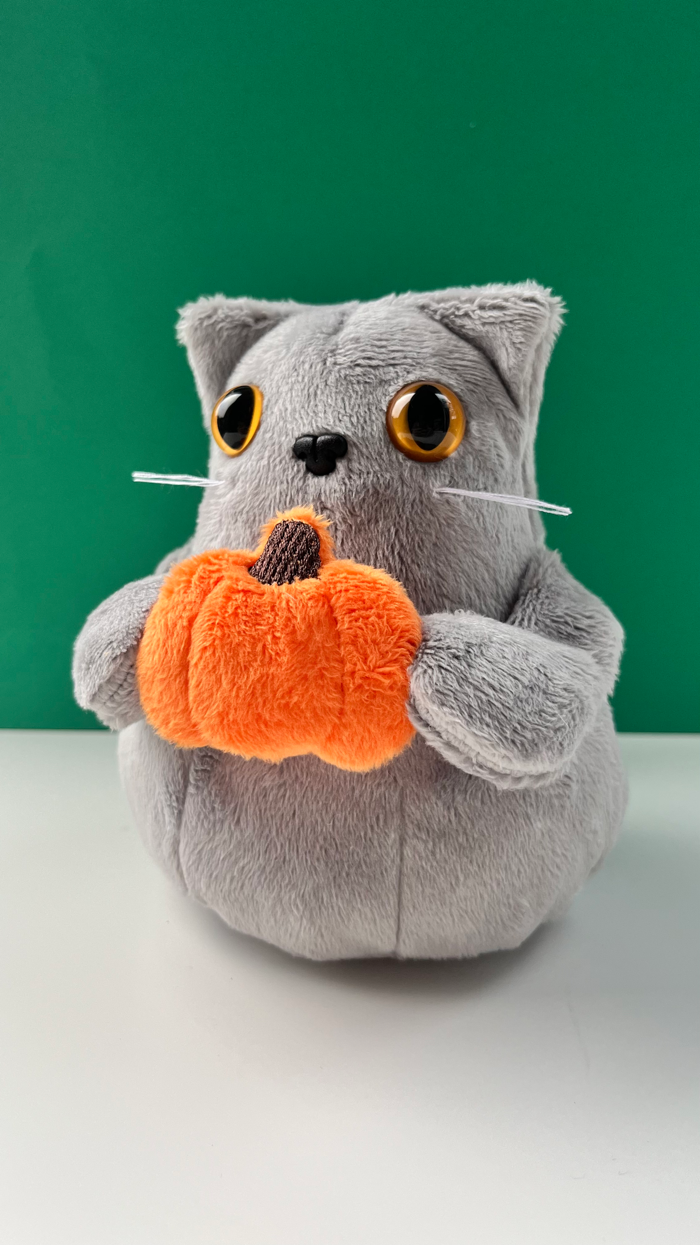 Grey cat plush holding a pumpkin accessory in front of a green background