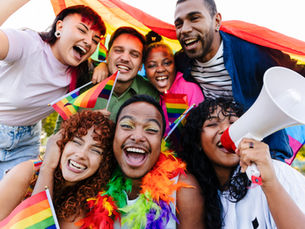 Diverse group of non-binary, gender-queer, and gay people celebrates with rainbow flags and a megaphone, wrapped in colorful banners, smiling joyfully outdoors.