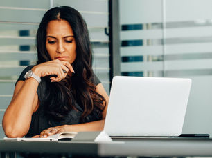 Gen X woman thinking with hand on chin, sitting at a desk with a laptop and notebook. Glass partition background, neutral colors, focused mood.