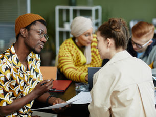 Group of four Gen Z people collaborating at a table with papers and laptops. One person wears a colorful shirt and beanie. Green background.