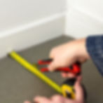 Hands measure a corner floor with a yellow tape measure and red utility knife. Gray coir backing being cut in place, white walls, blue sweater visible.