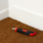 Red and black utility knife on brown coir matting near a white wall corner. The knife blade is exposed, set against the textured coconut coir.