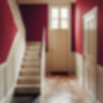 Staircase with white steps and railing, red walls, and a closed door under bright window in a warmly lit hallway. Checkerboard floor tiles.
