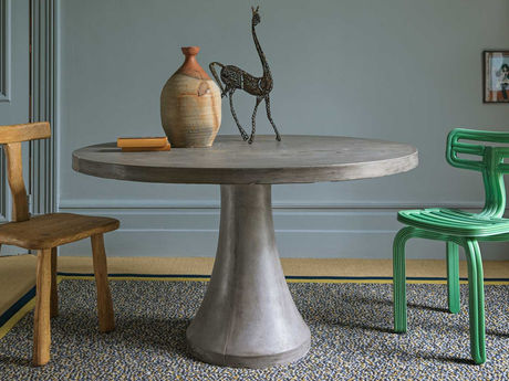 A round table with pottery and a metal sculpture is flanked by a wooden chair and a green chair on a patterned Collage Modernity Rug with Inner and outer wool felt borders in a muted room.