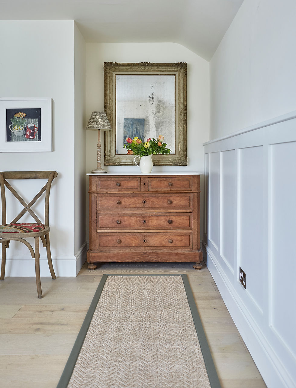 Bright hallway with wooden dresser, vase of flowers, large mirror, lamp, chair, and beige runner rug against white walls.