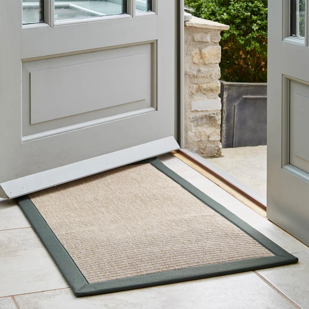 A gray door is partially open onto a tiled floor, revealing a beige mat with a dark border. A stone wall and greenery are visible outside.