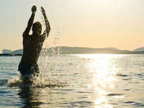 Lebensfreude wiederfinden: Eine Person steht bis zur Hüfte im Meerwasser, hebt die Arme nach oben, die Sonne spiegelt sich auf der Wasseroberfläche, im Hintergrund ist eine ruhige Küstenlinie zu sehen.