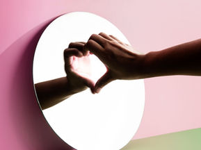 Selbstliebe, zwei Hände formen vor einem runden Spiegel ein Herz vor rosa Hintergrund mit grünem Farbverlauf