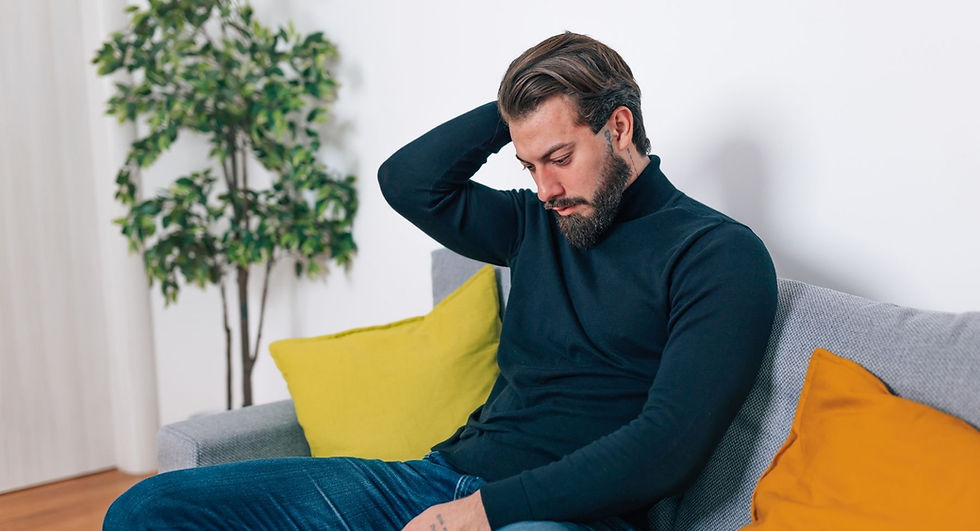 Gefühle: Mann sitzt nachdenklich auf einem Sofa, blickt nach unten und hält eine Hand an den Hinterkopf – ein Bild für innere Anspannung, Grübeln und den Umgang mit schwierigen Gefühlen.