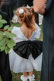 Flower girl wearing a white dress with a large black bow during an outdoor wedding ceremony at The Oaks at Plum Creek.