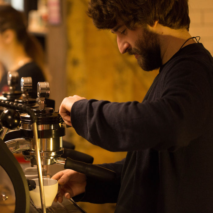 A man dressed in a black jumper uses a professional barista machine, coffee is pouring out into a cup. 

