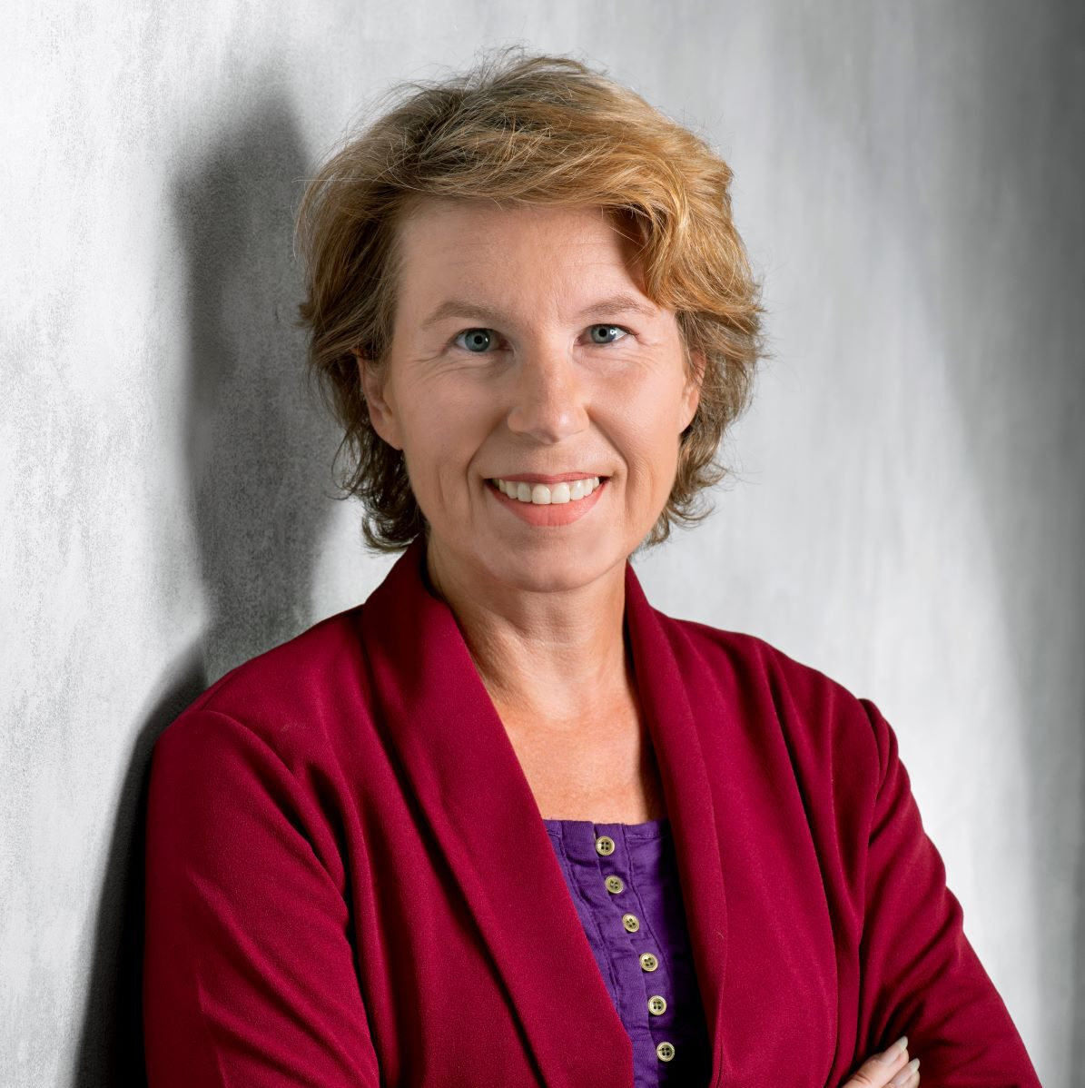 Sabine Hossenfelder, physicist and science communicator, smiling in a red jacket stands against a gray wall, arms crossed. Casual yet professional atmosphere, with a calm expression.