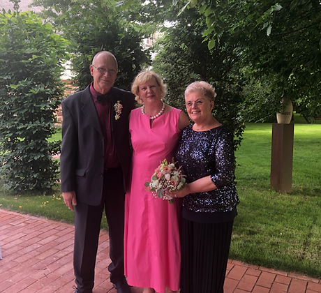 Ein älteres Paar, das Goldenen Hochzeit feiert. Sie sind festlich gekleidet, die Dame trägt einen Brautstrauß. Zwischem dem Paar steht eine blonde Frau im pinkfarbenen langen Kleid mit Perlenkette.
