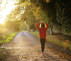 Woman walking down gravel pathway with hands on top of her head as though in despair or deep thought. Sun filtering through trees in front of the woman.