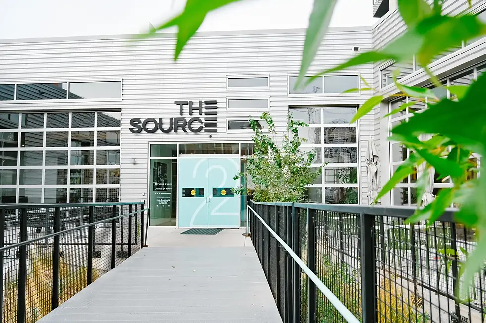 Modern white building entrance with THE SOURCE sign, aqua doors, black railings, and green leaves framing the walkway.