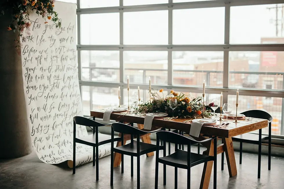 Elegant dining table set with candles and flowers by large windows; a handwritten calligraphy backdrop hangs at left.