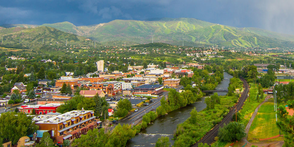 A mountain town beside a river and railroad, with green hills under dark storm clouds, bright buildings, and a moody skyline.