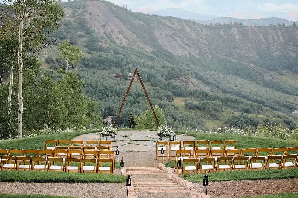 Outdoor mountain wedding setup with wooden chairs, a triangular arch, lanterns, and flowers on a green hillside under a calm sky
