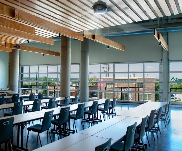 Empty modern classroom with long white tables and black chairs, tall windows, concrete columns, and wood ceiling beams in daylight