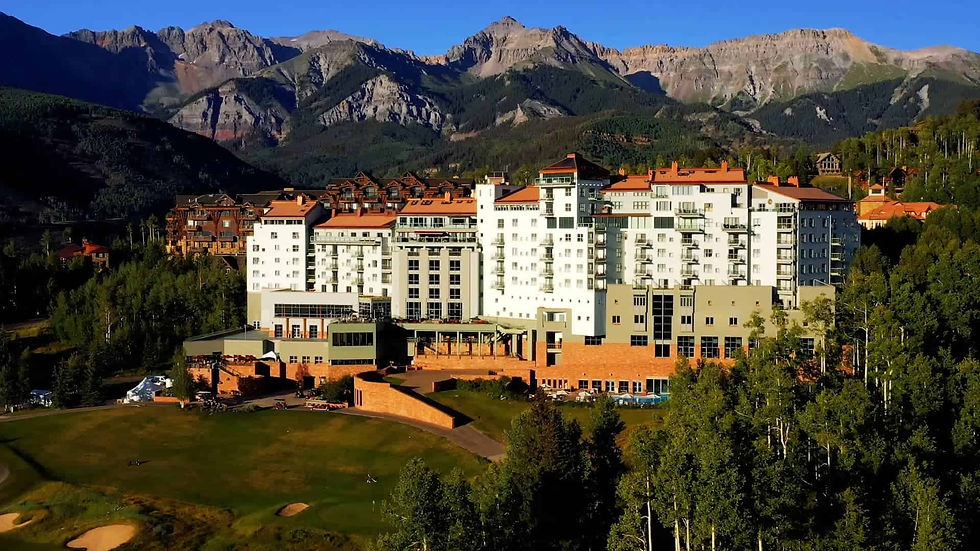 Mountain resort hotel beside a green golf course, framed by tall rocky peaks under a clear blue sky.