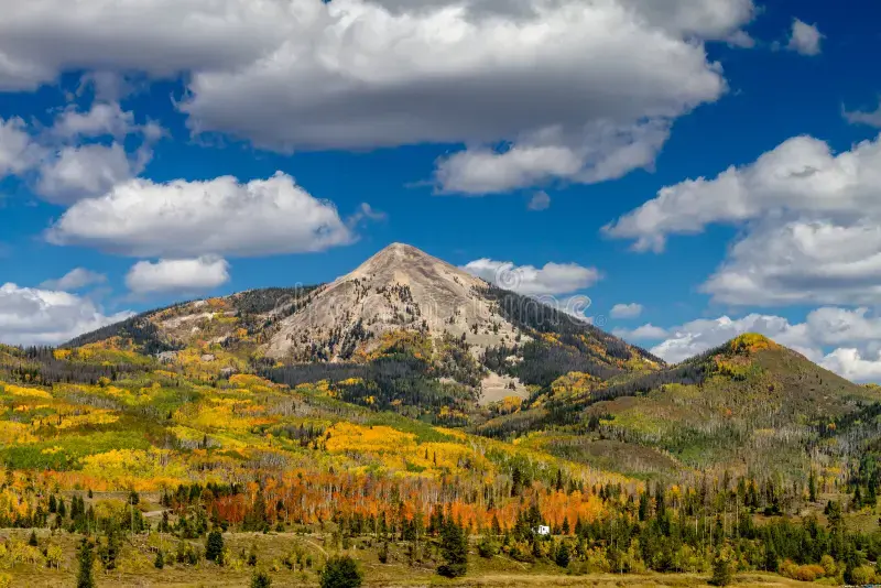 Mountain peak above colorful autumn forest under a blue sky with scattered white clouds.