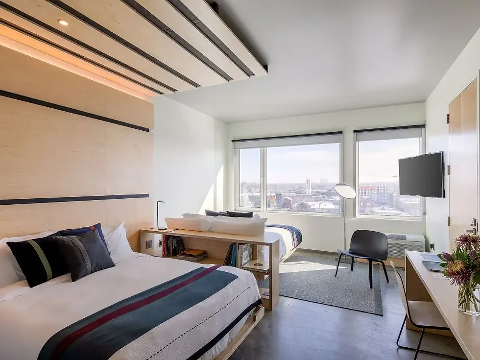Bright modern bedroom with two beds, large city-view windows, black chair, and desk in a minimalist white and wood room.