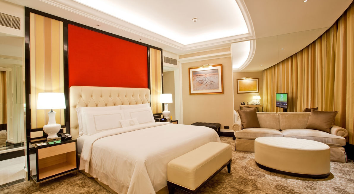 Large luxury hotel suite living room featuring a long beige sofa, a patterned area rug, a glass coffee table, a television, and a dining table in the background.