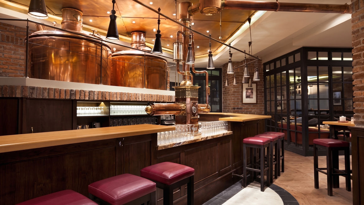 Classic restaurant bar featuring massive copper brewing tanks behind a wooden bar counter lined with red upholstered bar stools.
