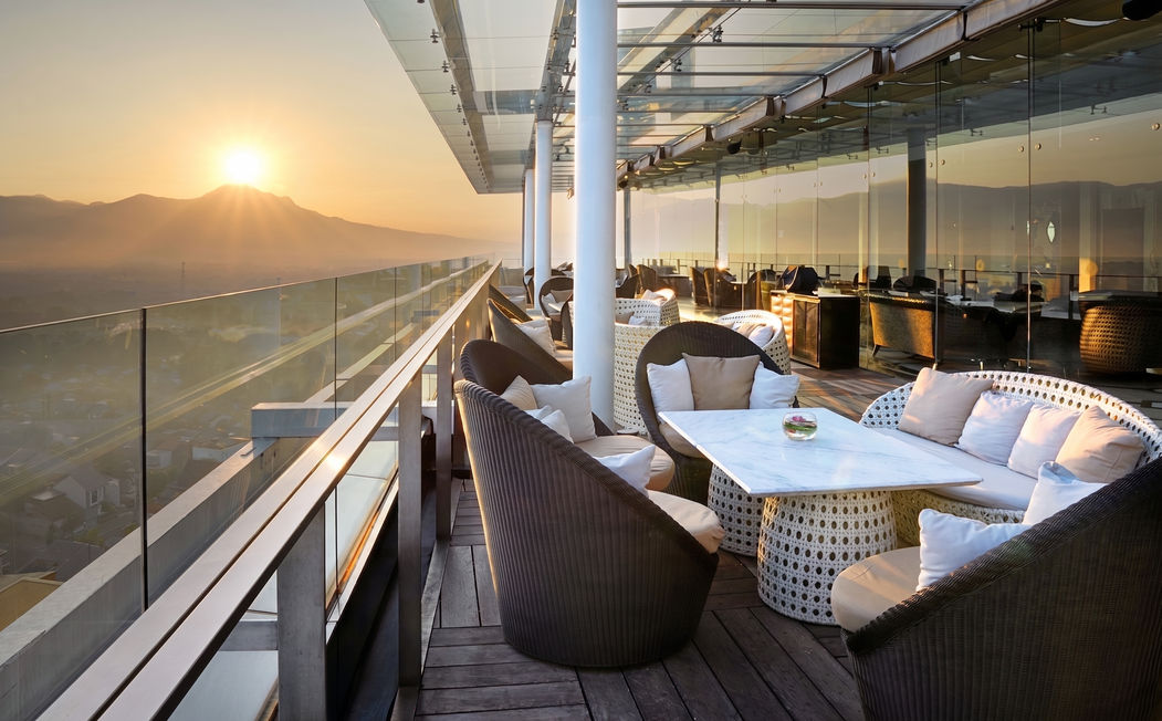 Outdoor rooftop hotel terrace at sunset featuring stylish wicker chairs, white tables, a glass railing, and a scenic view of the city and mountains in the distance.