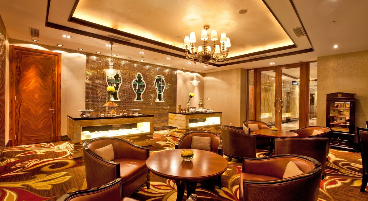 Dimly lit luxury hotel lounge with patterned carpets, leather armchairs arranged around round tables, and decorative vases on a back counter.