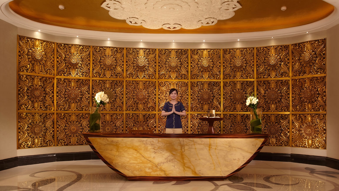 Hotel receptionist standing behind a glowing marble front desk, framed by a highly detailed, ornate golden patterned wall and warm ceiling lighting.