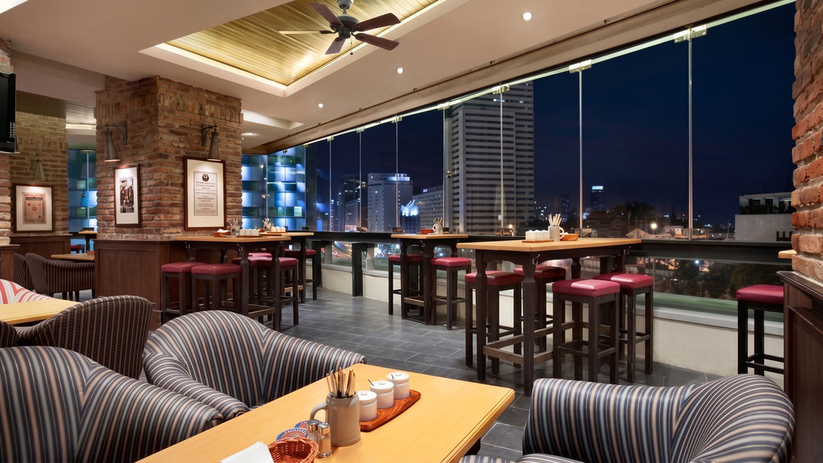 Restaurant seating area with large glass windows showing a city skyline at dusk, featuring striped lounge chairs in the foreground and high bar tables in the back.