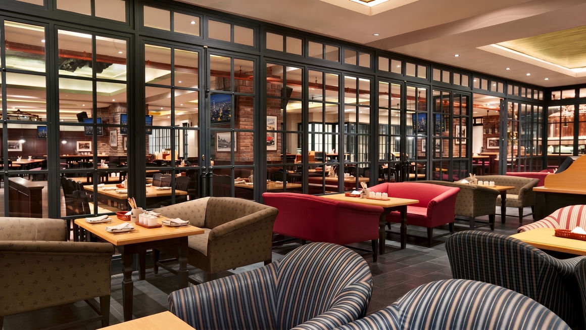 Restaurant interior showing striped armchairs, wooden tables, red sofa seating, and a large black-framed glass wall dividing the space.