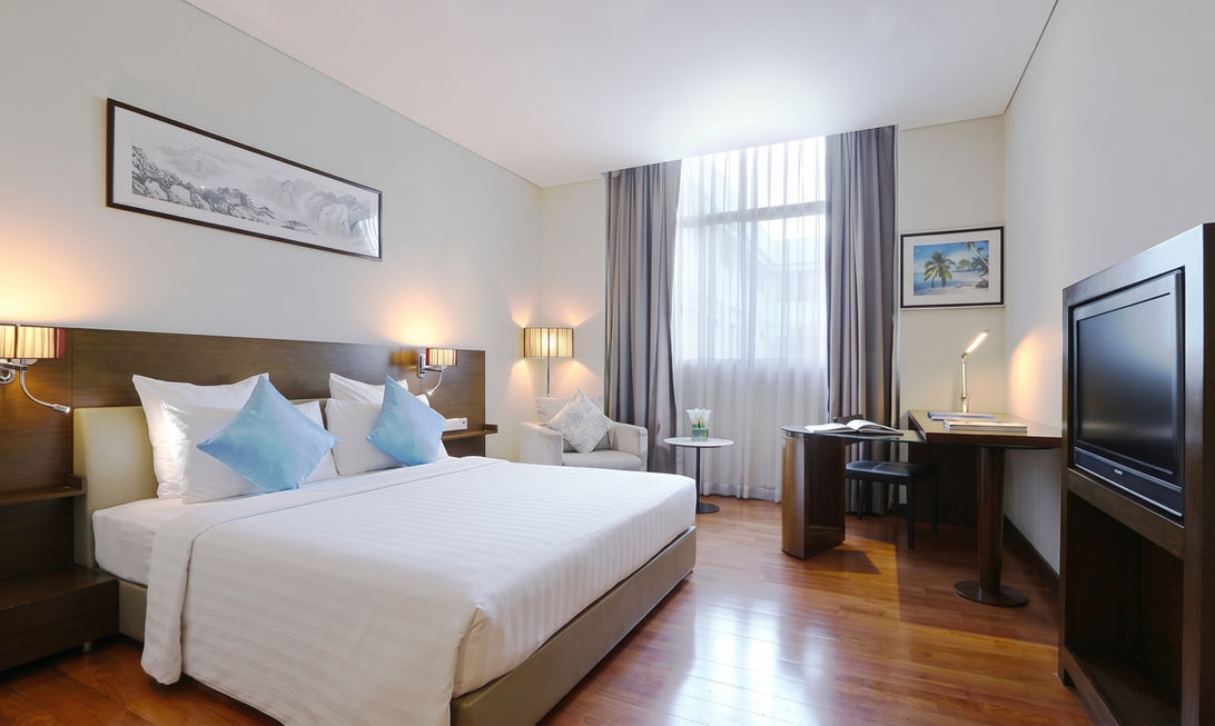 Dimly lit hotel bedroom interior with a large bed, blue pillows, and a window in the background.