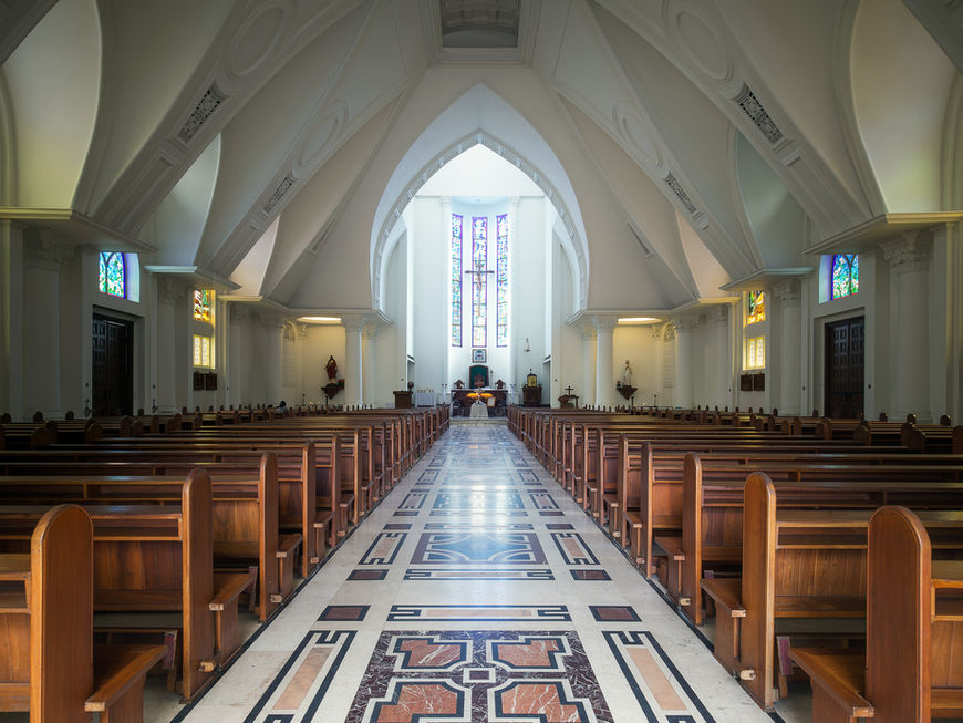 Central perspective down the main aisle of a church nave with patterned marble flooring, wooden pews on both sides, and high white arches leading to an altar with vertical stained-glass windows.
