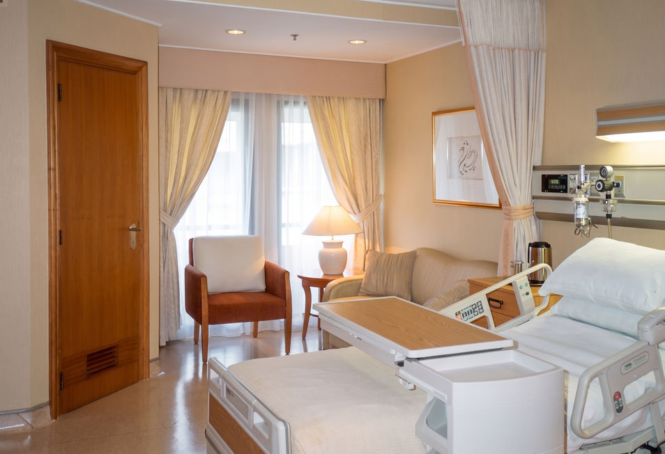 Hospital room interior showing a patient bed with an overbed table, a comfortable armchair, and large windows with sheer curtains.