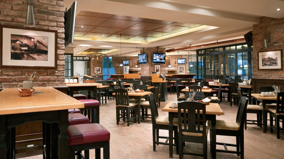 Large, warmly lit restaurant dining area featuring wooden tables, dark chairs, exposed brick columns, and framed pictures on the walls.