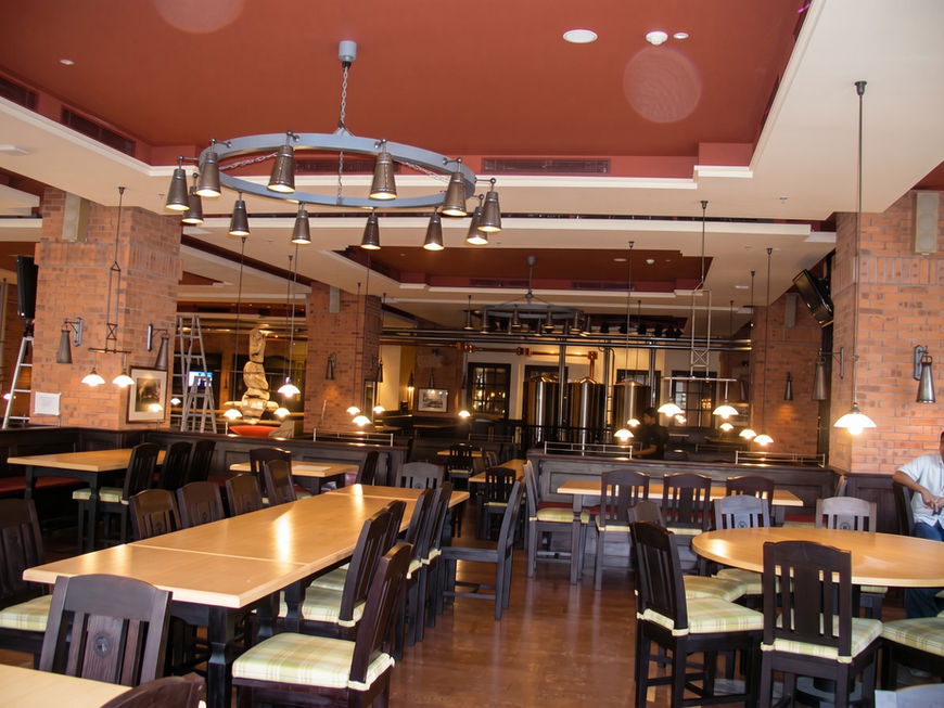 Spacious dining hall with long wooden communal tables, brick walls, and a large rustic ring chandelier hanging from the ceiling.