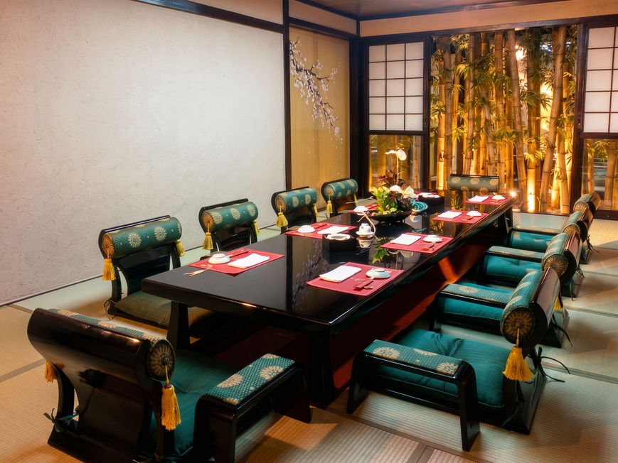Japanese private dining room with a long black table, green zaisu chairs on tatami mats, and bamboo decorations visible through the window.