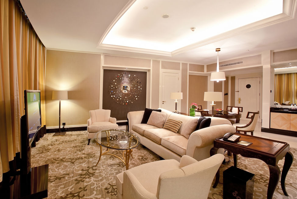 Large luxury hotel suite living room featuring a long beige sofa, a patterned area rug, a glass coffee table, a television, and a dining table in the background.