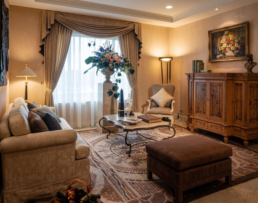 Interior view of a warmly lit hotel suite living area with a large sofa, a cushioned ottoman, dark wood furniture, and curtains.