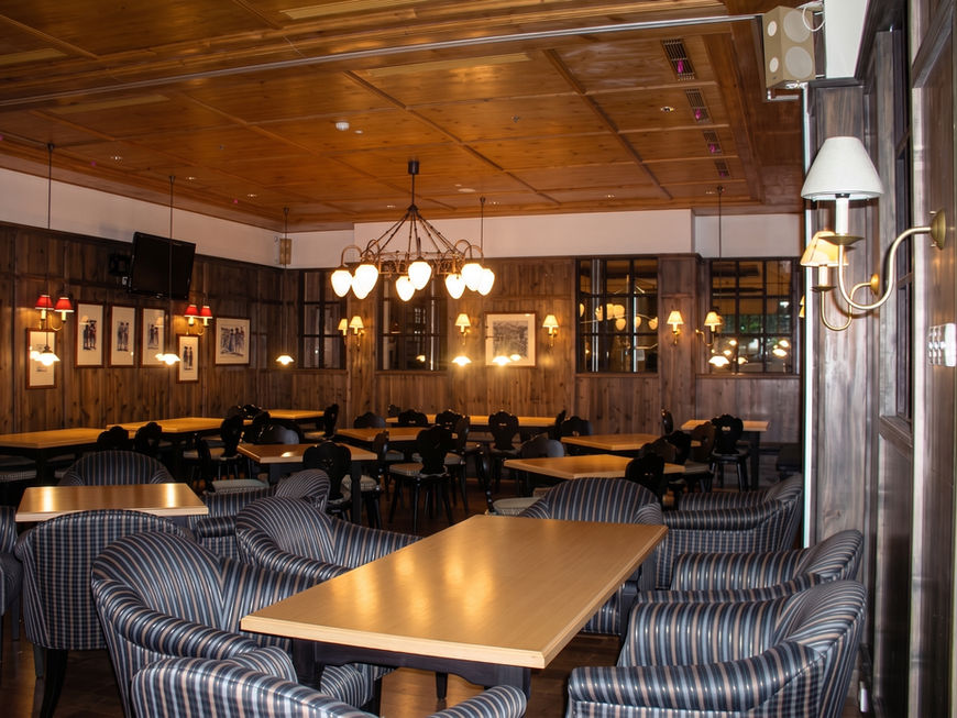 Restaurant dining room featuring a wooden ceiling, long dining tables, striped upholstered chairs, and ambient hanging lights.