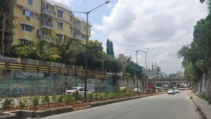 "Wide-angle view of Benson Town, Bengaluru, showing a mix of colonial-era architecture and modern buildings. The streets are lined with lush green trees, creating a serene and shaded environment. A few vehicles and pedestrians can be seen navigating the calm neighborhood, reflecting its residential charm."